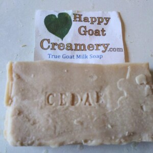 May include: A rectangular bar of goat milk soap with the word "CEDAR" stamped into it. A paper label with a green heart and the text "Happy Goat Creamery.com True Goat Milk Soap" is behind the soap.