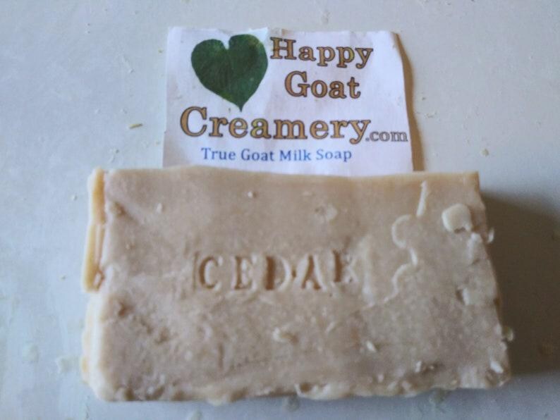 May include: A rectangular bar of goat milk soap with the word "CEDAR" stamped into it. A paper label with a green heart and the text "Happy Goat Creamery.com True Goat Milk Soap" is behind the soap.