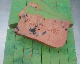 Può includere: Una pila di saponette rettangolari con esterno verde e interno rosa con macchie scure. La saponetta superiore presenta la parola Watermelon in rilievo, mostrando un design a strati che imita l'aspetto di un frutto tagliato a fette.