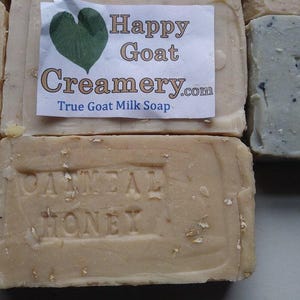 May include: Close-up of several bars of soap. One bar has the text "Oatmeal Honey" embossed on it. A label on another bar reads "Happy Goat Creamery.com True Goat Milk Soap" with a green heart graphic. The soaps are beige and light blue.