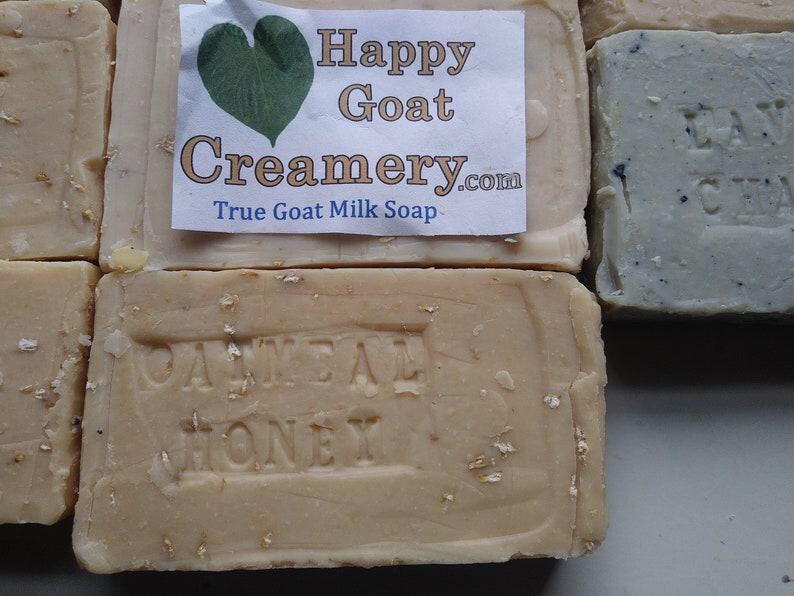 May include: Close-up of several bars of soap. One bar has the text "Oatmeal Honey" embossed on it. A label on another bar reads "Happy Goat Creamery.com True Goat Milk Soap" with a green heart graphic. The soaps are beige and light blue.