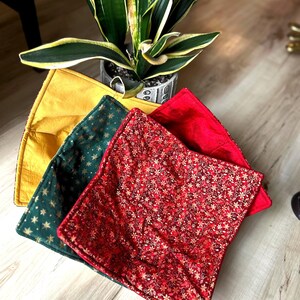 Op de afbeelding: Vier decoratieve vierkante kussens in geel, groen en rood. Het groene kussen heeft een gouden sterrenpatroon, terwijl het rode kussen een gouden bloemenontwerp heeft. Een potplant met groene en gele bladeren staat op de achtergrond.