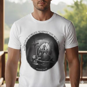 May include: White t-shirt featuring a black and white graphic of a shield, sword, and axe. The graphic is encircled by the text "NO WEAPON FORGED AGAINST YOU WILL PREVAIL" and "ISAIAH 54:17".