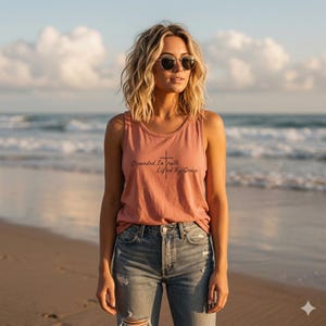 May include: A dusty rose sleeveless tank top featuring a black graphic print of a cross and the text Grounded In Truth Lifted By Grace, paired with light blue distressed denim jeans. The top has a round neckline and a relaxed fit.
