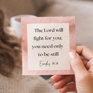 May include: A square card with a pink border and a white centre, featuring the text "The Lord will fight for you; you need only to be still Exodus 14:14" in black script. The card is held by a person.