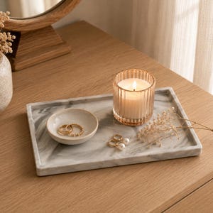 Op de afbeelding: Een rechthoekig dienblad van wit en grijs marmer met een klein beige keramieken schaaltje met twee gouden ringen, een geribbelde glazen kaarsenhouder met een brandende witte kaars en een takje gedroogde witte bloemen op het oppervlak.