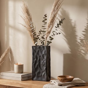 May include: A tall, rectangular black vase with a textured, uneven surface holds several dried pampas grass plumes and eucalyptus stems. Beside it sits a small, lit beige candle on two stacked books and a small wooden bowl on a linen cloth.