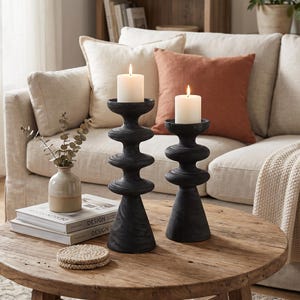 May include: Two black, tiered, sculptural candle holders with a matte finish, each topped with a lit white pillar candle. The holders feature a stacked, rounded design and are positioned on a round wooden table next to a small ceramic vase.