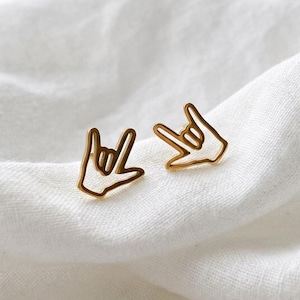 May include: A pair of gold-colored stud earrings in the shape of the "I love you" hand sign. The earrings are displayed on a white, textured fabric, with soft lighting.