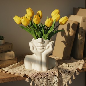 May include: A white ceramic face vase holding a bouquet of yellow tulips. The vase has a minimalist design with the face resting on its hands. The flowers are vibrant against the white vase. The vase sits on a lace doily on a wooden shelf.
