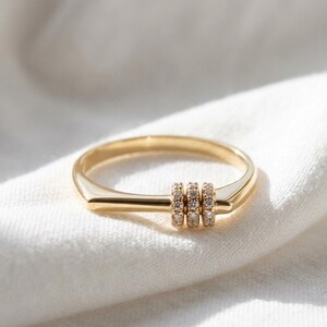Op de afbeelding: Een gouden ring met drie kleine, met diamanten bezette bandjes. De ring heeft een strak, modern design met een rechthoekige band die taps toeloopt naar een afgeronde vorm. De ring wordt gepresenteerd op een witte, getextureerde stof.