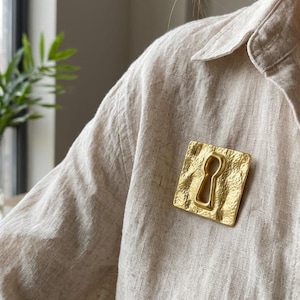 May include: A gold-colored keyhole brooch with a textured surface is pinned to a beige shirt. The brooch is square-shaped with a keyhole cutout in the center. The background includes a window and greenery.