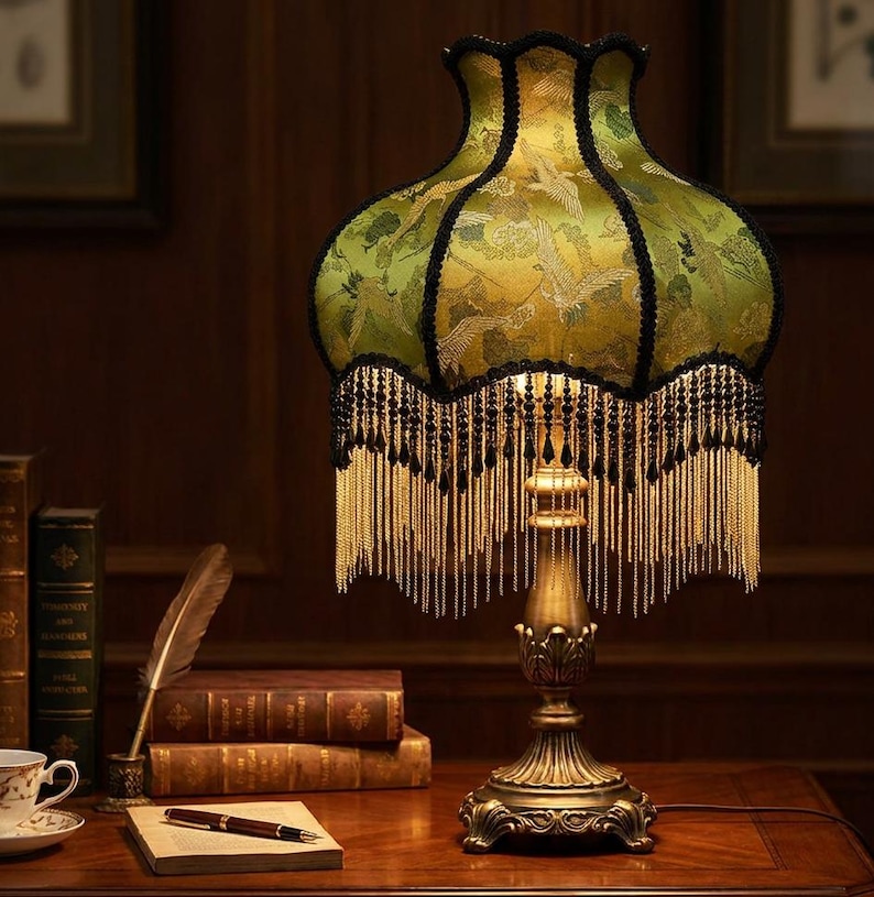 May include: A table lamp with a scalloped green fabric shade featuring a crane pattern, black trim, and long gold fringe. The lamp has a metallic, ornate base with a pedestal design. The light emits a warm glow from beneath the decorative shade.