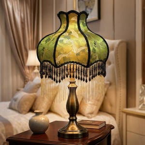 May include: A table lamp with a brass-toned metal base and a bell-shaped lampshade in lime green fabric. The shade features a pattern of white cranes and clouds, accented with black trim and a fringe of black beads and thin gold chains.