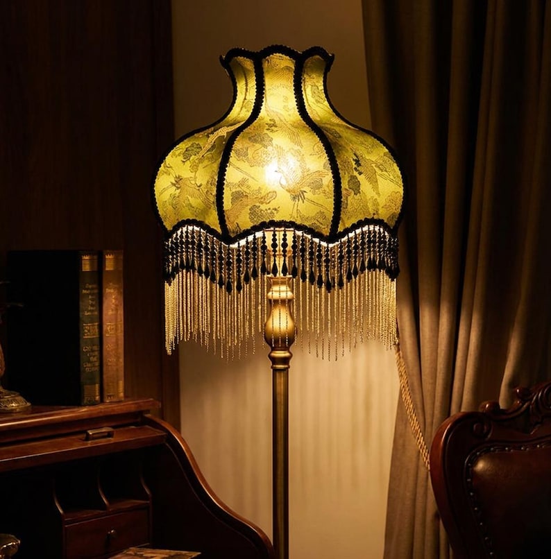 May include: A floor lamp featuring a bell-shaped, light green fabric shade with a dark bird and floral pattern. The shade is trimmed with black scalloped edges and long, dangling black and clear beaded fringe. It stands on a slender metal pole.