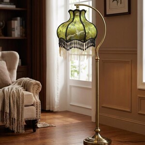 May include: A floor lamp with a curved brass-toned metal stand and a circular base. The lampshade is bell-shaped, featuring a light green fabric with a white crane pattern, finished with black trim and a long, layered fringe of black and white beads.