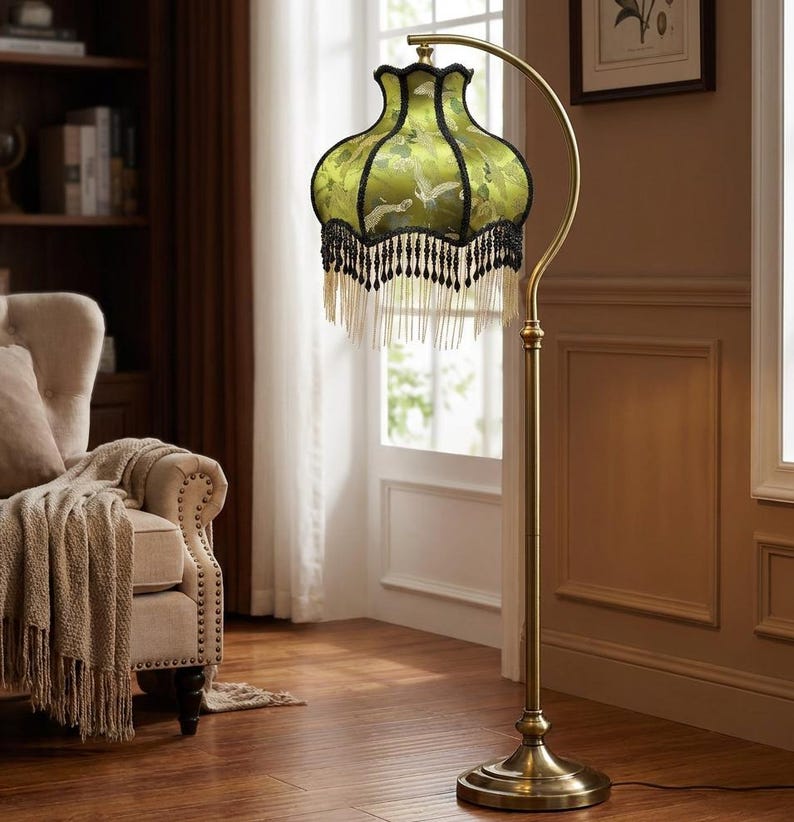 May include: A floor lamp with a curved brass-toned metal stand and a circular base. The lampshade is bell-shaped, featuring a light green fabric with a white crane pattern, finished with black trim and a long, layered fringe of black and white beads.