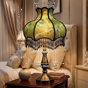 May include: A table lamp with a green fabric shade featuring a crane pattern, black trim, and hanging black and gold fringe. The lamp has a metallic base and measures 5.9 inches wide at the top, 13.8 inches wide at the base, and 14.5 inches tall.