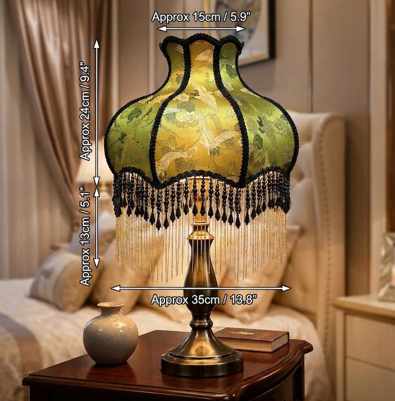 May include: A table lamp with a green fabric shade featuring a crane pattern, black trim, and hanging black and gold fringe. The lamp has a metallic base and measures 5.9 inches wide at the top, 13.8 inches wide at the base, and 14.5 inches tall.