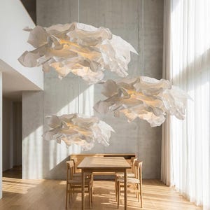 May include: Three white pendant lights with layered, ruffled, paper-like shades suspended from the ceiling. Each light fixture features a soft, glowing interior, creating a cloud-like appearance above a wooden dining table and chairs.