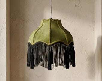 May include: A bell-shaped pendant lamp shade featuring a soft olive green fabric exterior with vertical stitched seams and a scalloped top edge. The bottom border is adorned with a long, dense black fringe trim consisting of thin hanging threads.