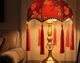 May include: A table lamp featuring a dome-shaped shade in vibrant red fabric with a gold floral pattern. The shade is trimmed with long, thin gold beaded fringe and red tassels. The lamp has a metallic gold stand with a clear crystal accent.