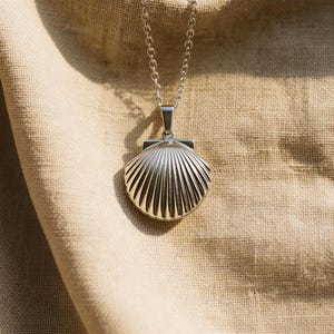 May include: A silver seashell pendant necklace. The pendant is a detailed, three-dimensional scallop shell design. The necklace has a delicate chain and a simple clasp. The background is a textured, tan fabric.