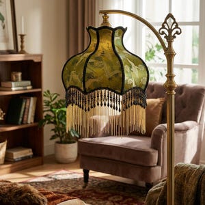 May include: A floor lamp with a curved brass stand and a scalloped fabric lampshade in olive green. The shade features a repeating crane pattern, black trim, and a decorative fringe made of black beads and long, thin gold metallic chains.