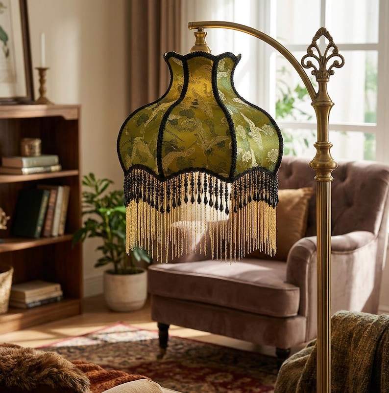 May include: A floor lamp with a curved brass stand and a scalloped fabric lampshade in olive green. The shade features a repeating crane pattern, black trim, and a decorative fringe made of black beads and long, thin gold metallic chains.