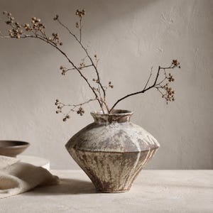 May include: A wide-bodied ceramic vase with a tapered base and a short, flared neck. The surface features a mottled, textured finish in shades of brown, cream, and white. A thin, bare branch with small, dried seed pods is placed inside the vase.