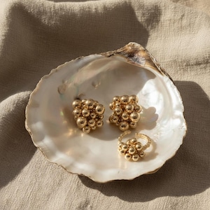 May include: Gold-colored jewelry set featuring a pair of stud earrings and a ring, each adorned with clustered spheres. The jewelry rests inside a large, iridescent seashell, set against a neutral fabric backdrop.