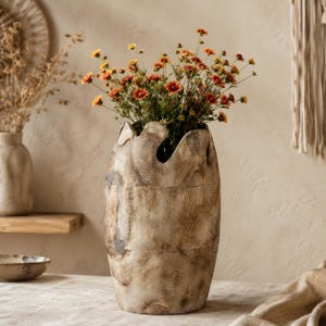 Puede incluir: Un jarrón cilíndrico de madera con un acabado natural en marrón claro desgastado y un borde superior irregular y dentado. El jarrón contiene un ramo vibrante de pequeñas flores de color naranja y amarillo con tallos y follaje verdes.