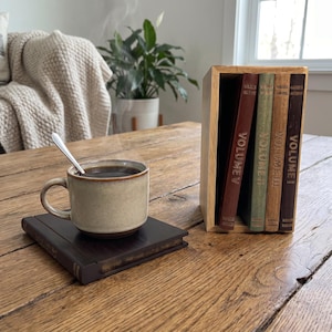 May include: A light-colored ceramic mug with a silver spoon sits on a dark brown book coaster. Beside it, a wooden book-shaped storage box holds four vertical books labeled Volume I, Volume II, Volume III, and Volume IV in gold lettering.