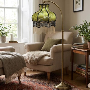 May include: A floor lamp with a curved brass stand and a bell-shaped lampshade. The shade is made of green fabric featuring a white crane pattern, finished with black trim and a long, layered fringe of black and cream-colored beads and tassels.