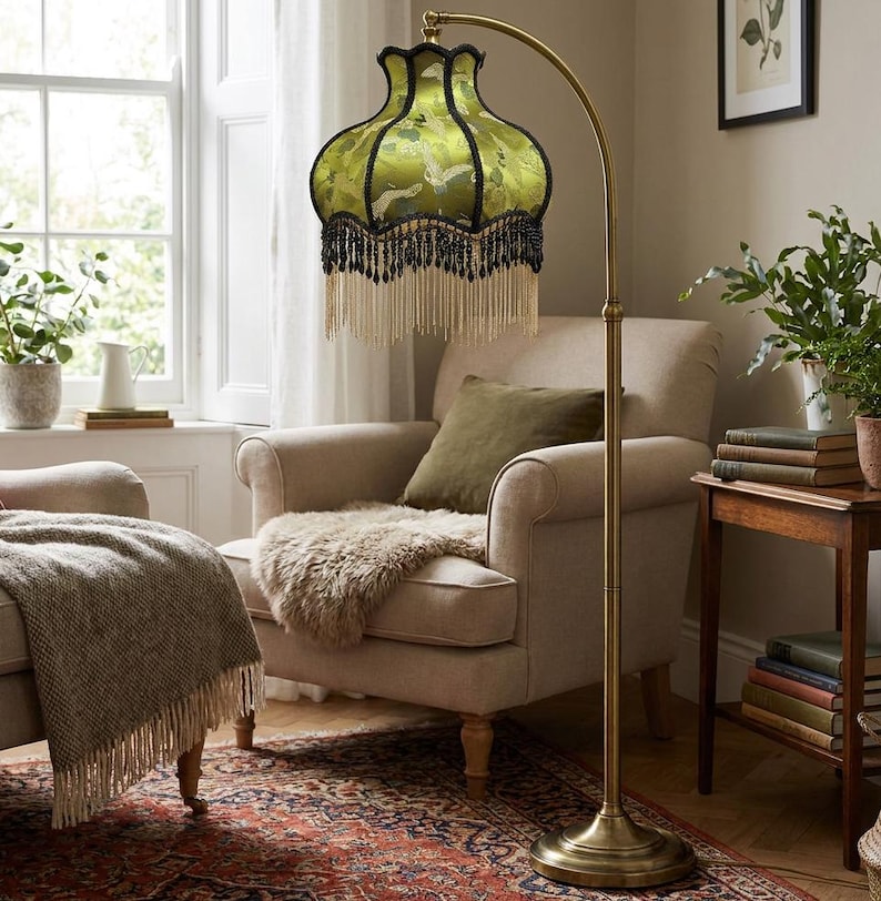 May include: A floor lamp with a curved brass stand and a bell-shaped lampshade. The shade is made of green fabric featuring a white crane pattern, finished with black trim and a long, layered fringe of black and cream-colored beads and tassels.