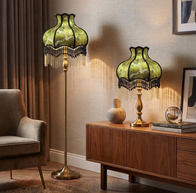 May include: A floor lamp and a matching table lamp, both featuring a brass-toned metal stand and a bell-shaped green fabric shade with a bird pattern, black trim, and long hanging black and gold fringe tassels around the bottom edge.