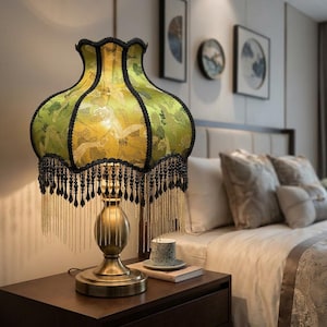 May include: A table lamp with a bell-shaped lime green fabric shade featuring a crane pattern, black scalloped trim, and long black and gold beaded fringe. The lamp has a metallic gold-toned pedestal base with a rounded, tiered design.