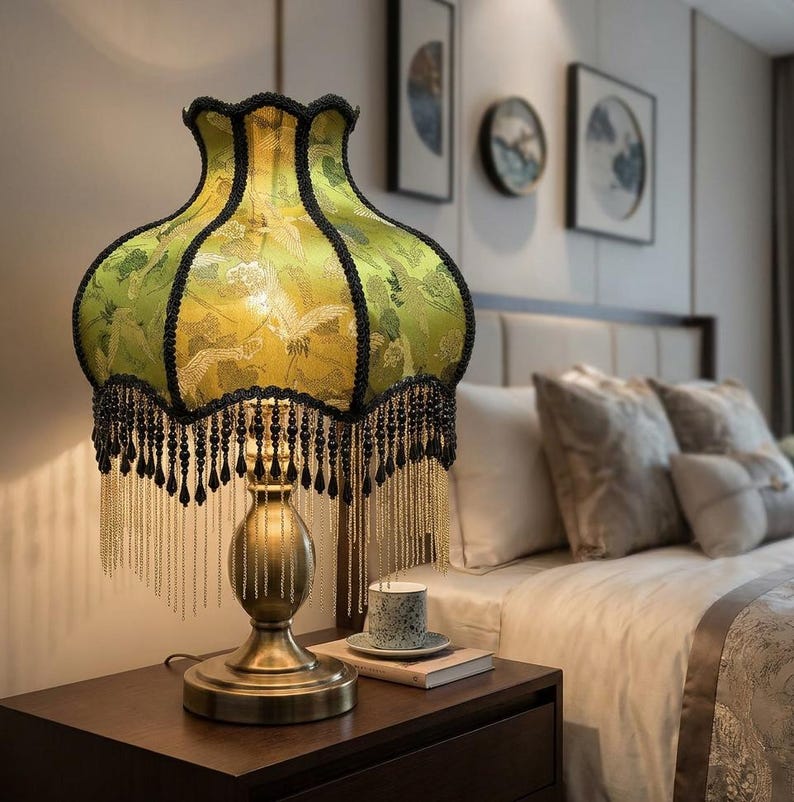 May include: A table lamp with a bell-shaped lime green fabric shade featuring a crane pattern, black scalloped trim, and long black and gold beaded fringe. The lamp has a metallic gold-toned pedestal base with a rounded, tiered design.