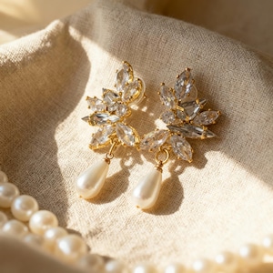 Puede incluir: Un par de pendientes dorados con brillantes cristales transparentes dispuestos en un diseño en forma de hoja. Cada pendiente presenta una perla de imitación en forma de lágrima. Los pendientes descansan sobre una tela beige, con un collar de perlas en primer plano.