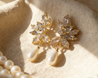 May include: A pair of gold-toned earrings with clear, sparkling crystal accents arranged in a leaf-like design. Each earring features a dangling, pear-shaped faux pearl. The earrings rest on a beige fabric, with a strand of pearls in the foreground.