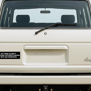 May include: A white SUV's rear with a black bumper and a bumper sticker. The sticker is black with a yellow smiley face and the text: "JUST TRYING TO HAVE A GOOD TIME DESPITE KNOWING FACTS AND INFORMATION."