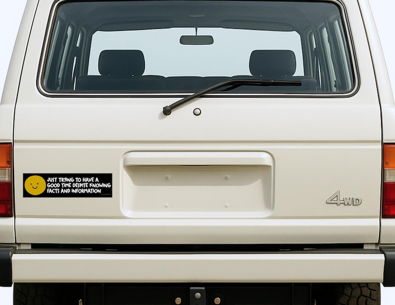 May include: A white SUV's rear with a black bumper and a bumper sticker. The sticker is black with a yellow smiley face and the text: "JUST TRYING TO HAVE A GOOD TIME DESPITE KNOWING FACTS AND INFORMATION."