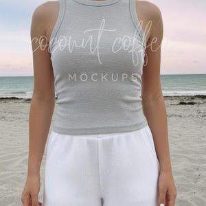 Puede incluir: Un top sin mangas de canalé gris claro y unos shorts blancos con textura de gofre. El texto "coconut coffee mockups" es visible en la parte superior. El atuendo se usa en una playa con un cielo rosa y azul.