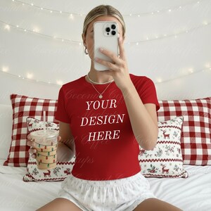 May include: A red t-shirt with the text "YOUR DESIGN HERE" in white. The person is holding a clear cup with a straw and a string of lights design. The background includes pillows with a red and white pattern.