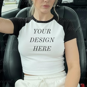 以下が含まれることがあります： 「YOUR DESIGN HERE」のテキストが入った白と黒のラグランクロップトップ。 シャツは黒い袖と白いボディです。 モデルは白いスウェットパンツを着用しています。
