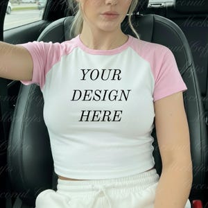 May include: White crop top with pink raglan sleeves. The shirt features the text "YOUR DESIGN HERE" in black, centered on the front. The model is wearing the shirt in a car.