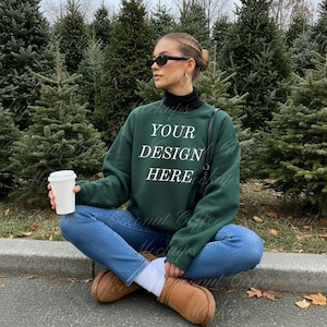 May include: A dark green sweatshirt with the text "YOUR DESIGN HERE" in white. The sweatshirt is worn with blue jeans and brown boots. A white coffee cup is held. The background features evergreen trees.