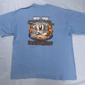 May include: Pale blue t-shirt with a cartoon image of the Tasmanian Devil on a sofa, holding a remote control. The text "CONTROL ISSUES" is above and below the image. The shirt has short sleeves.