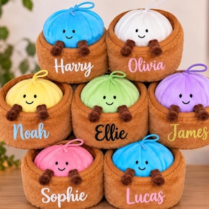 May include: A stack of seven plush toys, each featuring a colourful round character with a smiling face and brown dangling legs, nestled inside a brown textured basket. Each basket is personalised with a name in a unique script or print font.