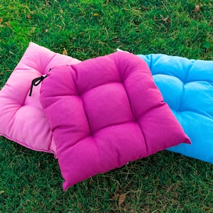 May include: Three square seat cushions in bright pink, magenta, and light blue. Each cushion features a tufted design with multiple stitched indentations and black fabric ties attached to the corners for securing the cushions to a chair.